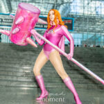 Cosplayer @pr1orities dressed as Atom Eve from Invincible at New York Comic Con 2025, holding a large pink spiked hammer and wearing a magenta bodysuit, cape, and boots while posing on the steps inside the Javits Center in NYC.