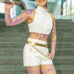 Cosplayer @longlivealex20 dressed as a K-Pop Demon Hunters character at New York Comic Con 2025, wearing a white and gold outfit with purple markings and purple hair, holding a sword over her shoulder at the Javits Center in NYC.