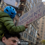 A child wearing a blue helmet and green jacket sits on an adult’s shoulders, holding a cardboard sign that reads “Why Is the President Mean?” during the No Kings March in New York City on October 18, 2025.