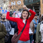 A young woman in a red shirt holds up a cardboard protest sign that reads “No Excuses, No Kings” while smiling during the No Kings March in New York City on October 18, 2025.
