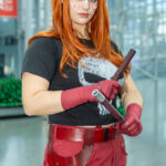 Cosplayer @magiciaansred dressed as a Daredevil and Punisher hybrid at New York Comic Con 2025, wearing a black Punisher T-shirt, red tactical pants, and red gloves while holding batons inside the Javits Center in NYC.