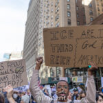Protesters at the No Kings March in New York City hold cardboard signs reading “ICE Are the New Third Reich, God Bless Amerikkka” and “If It’s About Criminals, Why Was One Elected??” surrounded by tall buildings and flags during the march from Times Square to 14th Street.