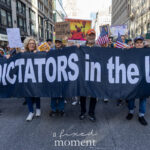 A large group of protesters marches through New York City holding a wide banner that reads “No Dictators in the USA” during the No Kings March on October 18, 2025.