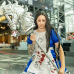 Cosplayer @sarahwood.cos dressed as Alice from American McGee’s Alice at New York Comic Con 2025, wearing a blue dress and blood-spattered white apron while holding a large armored horse-head weapon inside the Javits Center in NYC.