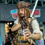 Cosplayer @cremsparrow dressed as Captain Jack Sparrow from Pirates of the Caribbean at New York Comic Con 2025, holding a drink and sword while posing inside the Javits Center in NYC.