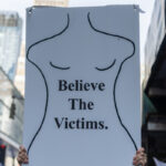 A protester at the No Kings March in New York City holds a sign with a simple black outline of a female body and the words “Believe The Victims.”