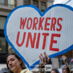A protester at the No Kings March in New York City holds a heart-shaped sign outlined in blue with bold red letters reading “Workers Unite.”