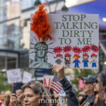 A protester at the No Kings March in New York City holds a handmade sign featuring the Statue of Liberty, red feathers, and the text “Stop Talking Dirty to Me.”