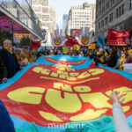 A crowd of protesters carries a large red and yellow banner reading “No Kings, No Cuts” during the No Kings March in New York City, surrounded by colorful signs and American flags.