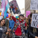 A group of young protesters holding colorful signs and flags march together at the No Kings March in New York City, with slogans including “No Kings No Fascists” and “Kick Your Local Racist.”