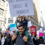 A man carries a child on his shoulders while the child holds a protest sign that reads “Only Butterflies Should Be Monarchs” during the No Kings March in New York City.