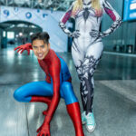 Cosplayers @spidey_ty and @oliviabettyrain dressed as Spider-Man and Symbiote Spider-Woman at New York Comic Con 2025, posing inside the Javits Center with Spider-Man crouched in action and Spider-Woman standing confidently beside him.