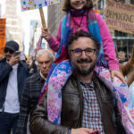 Father carrying daughter on shoulders during No Kings March in New York City. The young girl holds a colorful sign that reads “I know one thing… I don’t want a king!” amid a crowd of protesters.