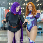 Two cosplayers from @hotnpcs dressed as Raven and Starfire from Teen Titans at New York Comic Con 2025, posing with their hands extended in dynamic stances under the bright lights of the Javits Center.