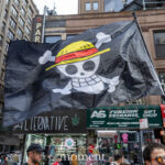 A black flag featuring the straw-hat skull logo from One Piece waves above protesters at the No Kings March in New York City.