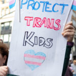 Protester at No Kings March in NYC holds a handmade sign that reads “PROTECT TRANS KIDS” with a heart in the colors of the transgender pride flag.