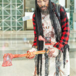 Cosplayer @artist_frankie dressed as a zombie lumberjack at New York Comic Con 2025, holding a bloody axe and wearing a red flannel shirt, apron, and black hat with dreadlocks at the Javits Center in NYC.