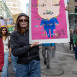 Protester wearing sunglasses and a hoodie holds a handmade collage poster of Donald Trump with ransom-note-style lettering during the No Kings March in NYC.