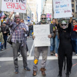 Three protesters dressed as zombies hold handmade political signs during the No Kings March in New York City, including messages like “EAT THE BRAINS” and “Trump Baaad!”