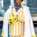 Cosplayer dressed as Admiral Kizaru from One Piece at New York Comic Con 2025, wearing a yellow striped suit, white admiral coat, and sunglasses while holding a glowing light prop outside the Javits Center in NYC.