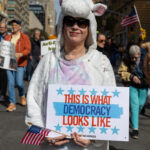 A person wearing a white unicorn costume holds a sign that says “THIS IS WHAT DEMOCRACY LOOKS LIKE” and a small American flag at the No Kings March in New York City.