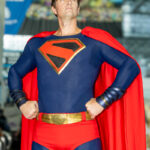 Cosplayer @rockyf150 dressed as an Art Deco–inspired Superman at New York Comic Con 2025, standing heroically with hands on hips, wearing a blue suit with a gold-trimmed red “S” emblem, red cape, and gold belt.