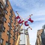 A colorful knitted protest sign reading “F*** ICE” attached to a wooden pole, held high against the backdrop of Manhattan buildings and blue sky during the No Kings March in New York City.