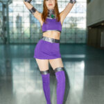 Cosplayer @hailee.1997 dressed as Starfire from Teen Titans at New York Comic Con 2025, wearing a purple and silver outfit with a green gemstone, posing with raised arms inside the Javits Center.