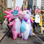 Two people wearing inflatable unicorn and axolotl costumes stand together at the No Kings March in New York City, holding a circular sign that reads “Amphifa – Amphibians Against Fascism.”