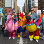 Three protesters wearing inflatable flamingo, rooster, and parrot costumes march down a Manhattan street during the No Kings March in New York City, holding flags and smiling.