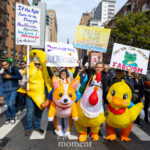 Protesters dressed in inflatable animal and fruit costumes hold signs advocating for democracy, peace, and justice during the No Kings March in New York City.
