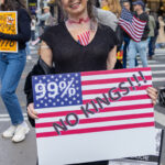 Woman in black dress and hat with a veil holds a red, white, and blue “99% NO KINGS!!!” protest sign at the No Kings March in New York City.