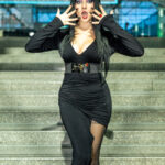 Cosplayer @dani_apples as Elvira, Mistress of the Dark, at New York Comic Con 2025, posing on the Javits Center steps in a black slit gown with dramatic makeup and black nails.