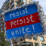 Blue protest sign with silver tinsel border reading “resist, persist, unite!” held up during the No Kings March in New York City.