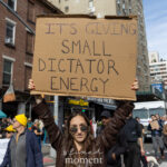 Protester wearing sunglasses and a brown jacket holds a cardboard sign reading “IT’S GIVING SMALL DICTATOR ENERGY” at the No Kings March in New York City.