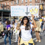 Woman smiling and holding a protest sign that reads “Resisting Bitch Face” with a crossed-out crown symbol during the No Kings March in New York City.