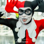 Cosplayer @motleycosplay as Harley Quinn in her red and black jester costume at New York Comic Con 2025, posing playfully with hands raised and tongue out inside the Javits Center.