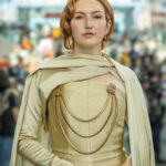 Cosplayer @laurendoescosplay dressed as Mon Mothma from Star Wars at New York Comic Con 2025, wearing a gold gown with ornate chain details and a matching cape, posing calmly in the Javits Center.