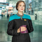Cosplayer @ella.s.christy dressed as Jadzia Dax from Star Trek: Deep Space Nine at New York Comic Con 2025, wearing a black and teal Starfleet uniform with Trill spots and holding a purple mug inside the Javits Center.