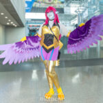 Cosplayer @heyghoulhey dressed as a fantasy harpy character with gold armor, purple and pink wings, yellow talon feet, and a red wig, posing with arms spread wide inside the Javits Center at New York Comic Con 2025.