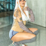 Cosplayer @goyarice dressed as Mirko from My Hero Academia crouches confidently, wearing a white wig, red glasses, beige corset, gray leg warmers, and iridescent platform heels inside the Javits Center at New York Comic Con 2025.