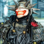 Cosplayer @critical.mask dressed as The Batman Who Laughs, wearing a spiked metal helmet, red grin mask, and detailed black leather armor with buckles and straps at New York Comic Con 2025 inside the Javits Center.
