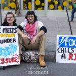 Two protesters sitting on a city curb with handmade signs reading “ICE melts faster under pressure” and “Fox News calls this a riot?” during the No Kings March in New York City.