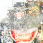 Cosplayers @critical.mask and @steampunkdbatmanvol2 dressed as The Batman Who Laughs and Steampunk Batman at New York Comic Con 2025, illuminated by bright light with reflective metal and gothic armor details inside the Javits Center.