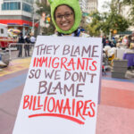 Smiling protester wearing a frog hat holds a sign that reads “They Blame Immigrants So We Don’t Blame Billionaires” during the No Kings March in New York City.