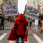 Protester in a red cloak holds two signs that read “Defend Democracy” and “We Will Not Be Silenced” during the No Kings March in New York City.