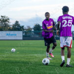Two soccer players from the Queens team wearing purple jerseys practice passing the ball on the field before “The Cost of Living Classic” at Maimonides Park in Coney Island.