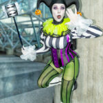 Cosplayer @_luna.foxx dressed as a Beetlejuice-inspired Harley Quinn, wearing a striped top, purple corset, green tights, and a jester hat with pumpkin charms, posing with a hammer inside the Javits Center at New York Comic Con 2025.