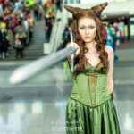Cosplayer @gennareading dressed as Jude Duarte from The Cruel Prince points a sword toward the camera while wearing a green corset gown with horn-styled hair inside the Javits Center at New York Comic Con 2025.