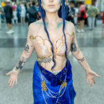 Cosplayer @gennareading dressed as Nicasia from The Cruel Prince stands with arms open at New York Comic Con 2025, wearing a pearl shell top with gold accents, blue braided hair, and a flowing blue skirt adorned with pearls.